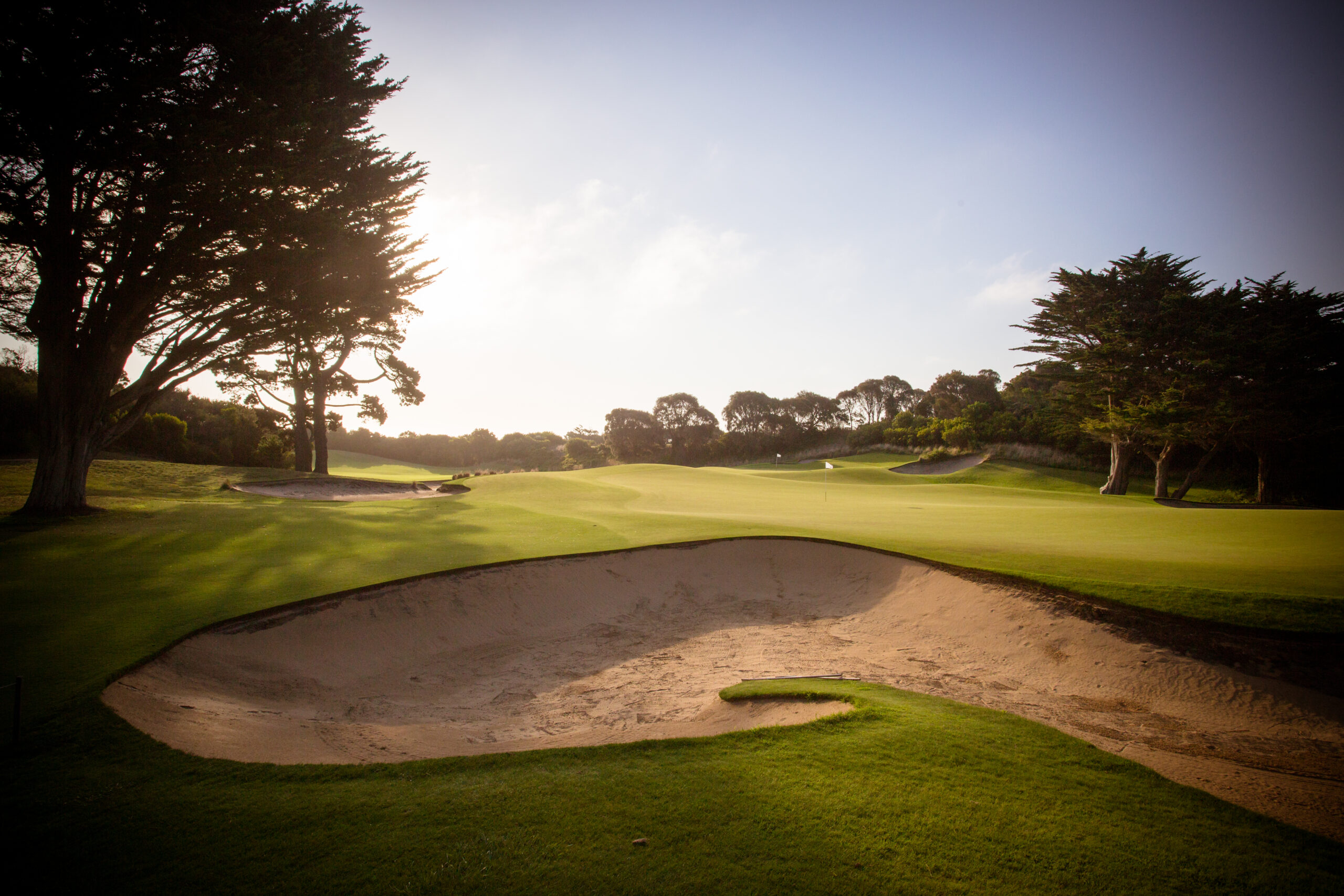 A golf course with a sand bunker, trees, and clear skies.