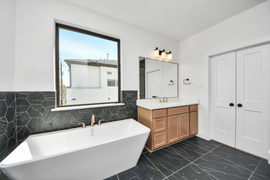 Modern bathroom with a freestanding tub, large window, wood vanity, and black tile floor.